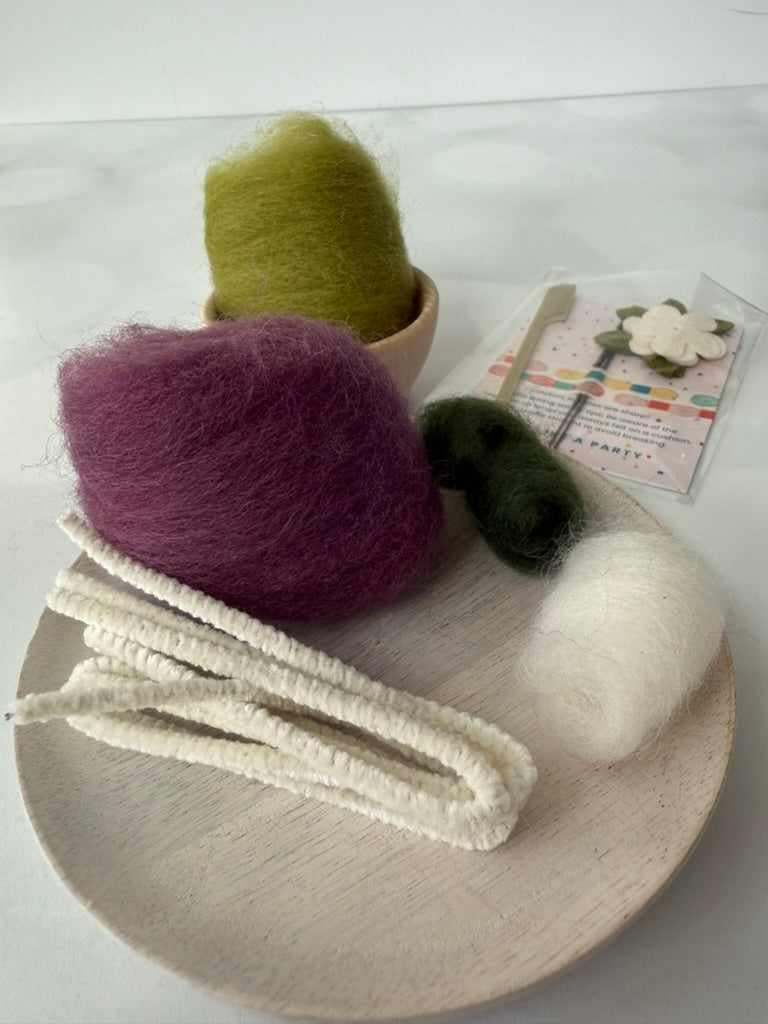 Needle felting kit including three balls of yarn in green, purple, and white on a wooden board with a color swatch in the background.