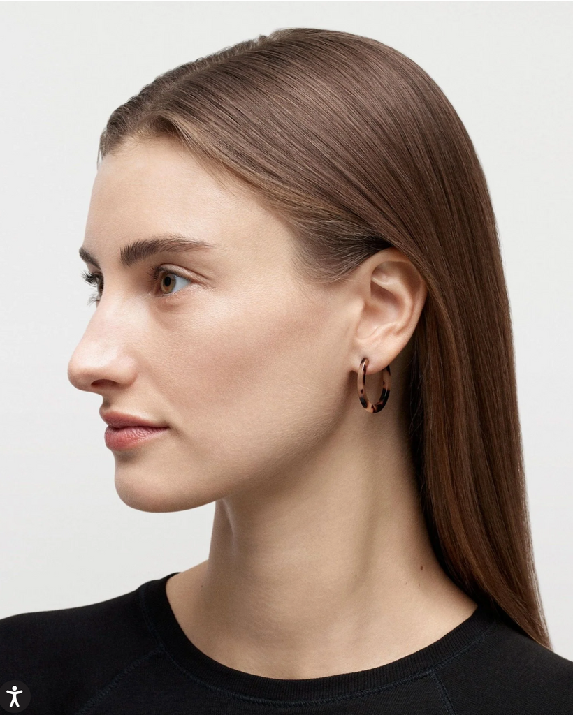 Woman with long brown hair wearing a black top against a plain background with tortoise shell hoop earrings.